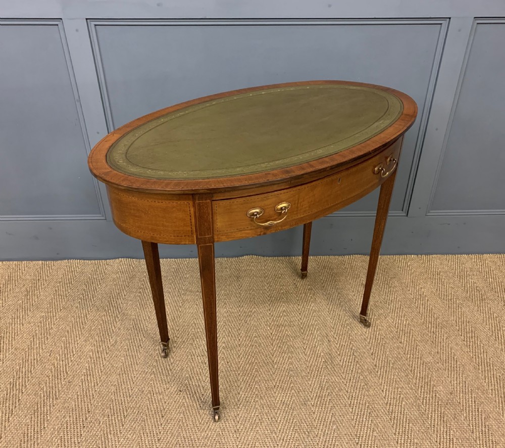 An Edwardian oval mahogany writing table featuring a green leather inset top, single frieze drawer with brass bail handles, and square tapered legs with castors. View VIII