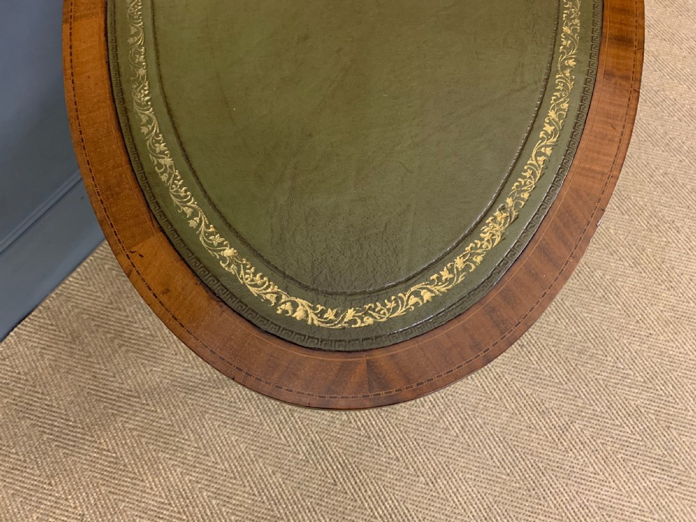 An Edwardian oval mahogany writing table featuring a green leather inset top, single frieze drawer with brass bail handles, and square tapered legs with castors. View IX