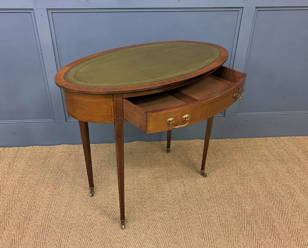 An Edwardian oval mahogany writing table featuring a green leather inset top, single frieze drawer with brass bail handles, and square tapered legs with castors. View XI
