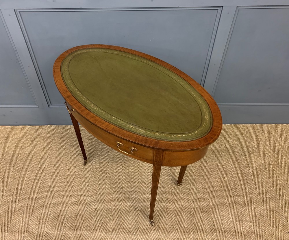 An Edwardian oval mahogany writing table featuring a green leather inset top, single frieze drawer with brass bail handles, and square tapered legs with castors. View XII