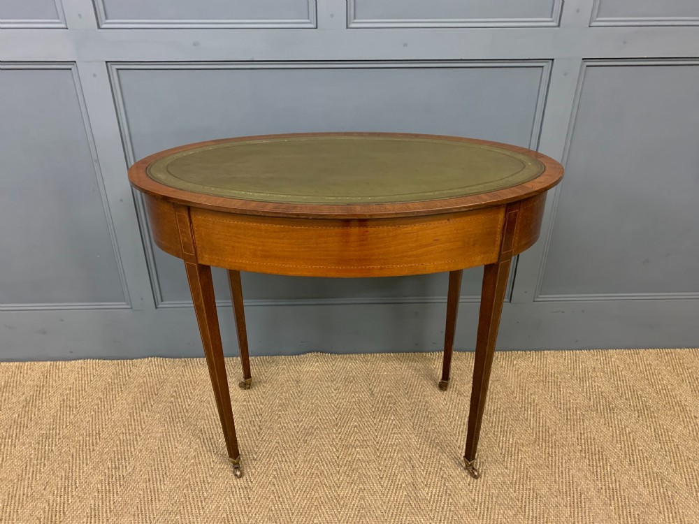 An Edwardian oval mahogany writing table featuring a green leather inset top, single frieze drawer with brass bail handles, and square tapered legs with castors. View XIII