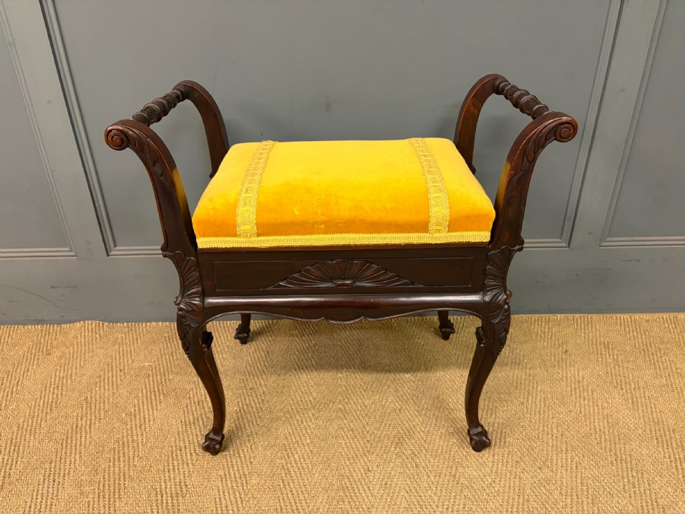 An antique dark mahogany piano stool featuring carved cabriole legs, scroll side handles, a carved shell apron motif, and a padded yellow velvet seat.