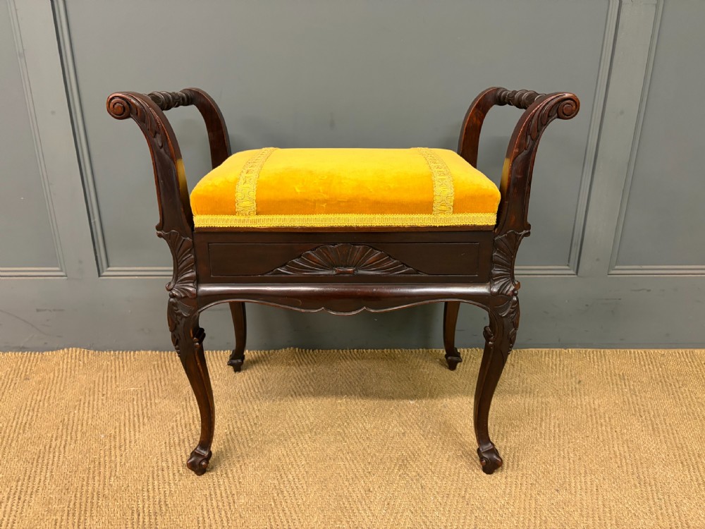 An antique dark mahogany piano stool featuring carved cabriole legs, scroll side handles, a carved shell apron motif, and a padded yellow velvet seat. View II