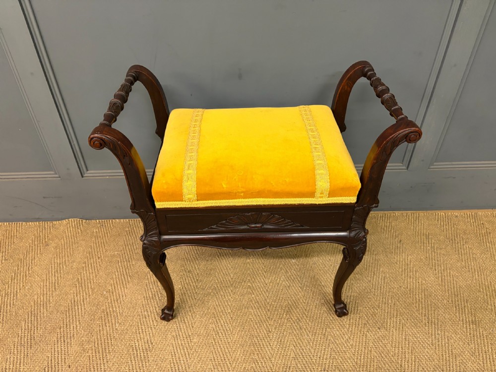 An antique dark mahogany piano stool featuring carved cabriole legs, scroll side handles, a carved shell apron motif, and a padded yellow velvet seat. View III