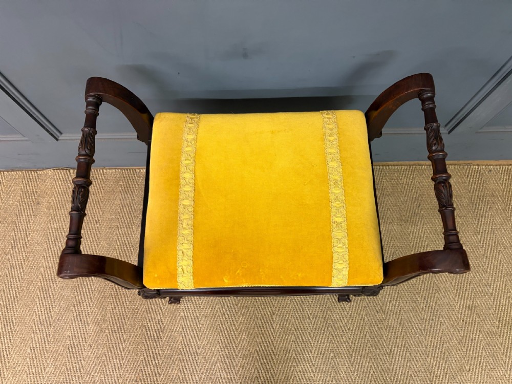 An antique dark mahogany piano stool featuring carved cabriole legs, scroll side handles, a carved shell apron motif, and a padded yellow velvet seat. View IV