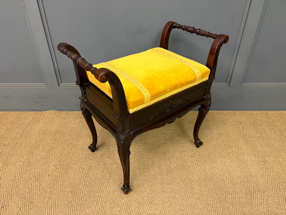 An antique dark mahogany piano stool featuring carved cabriole legs, scroll side handles, a carved shell apron motif, and a padded yellow velvet seat. View V