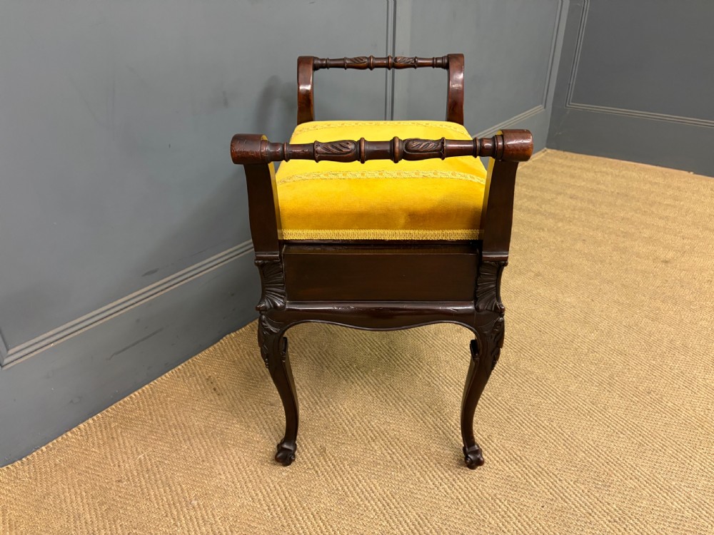 An antique dark mahogany piano stool featuring carved cabriole legs, scroll side handles, a carved shell apron motif, and a padded yellow velvet seat. View VI