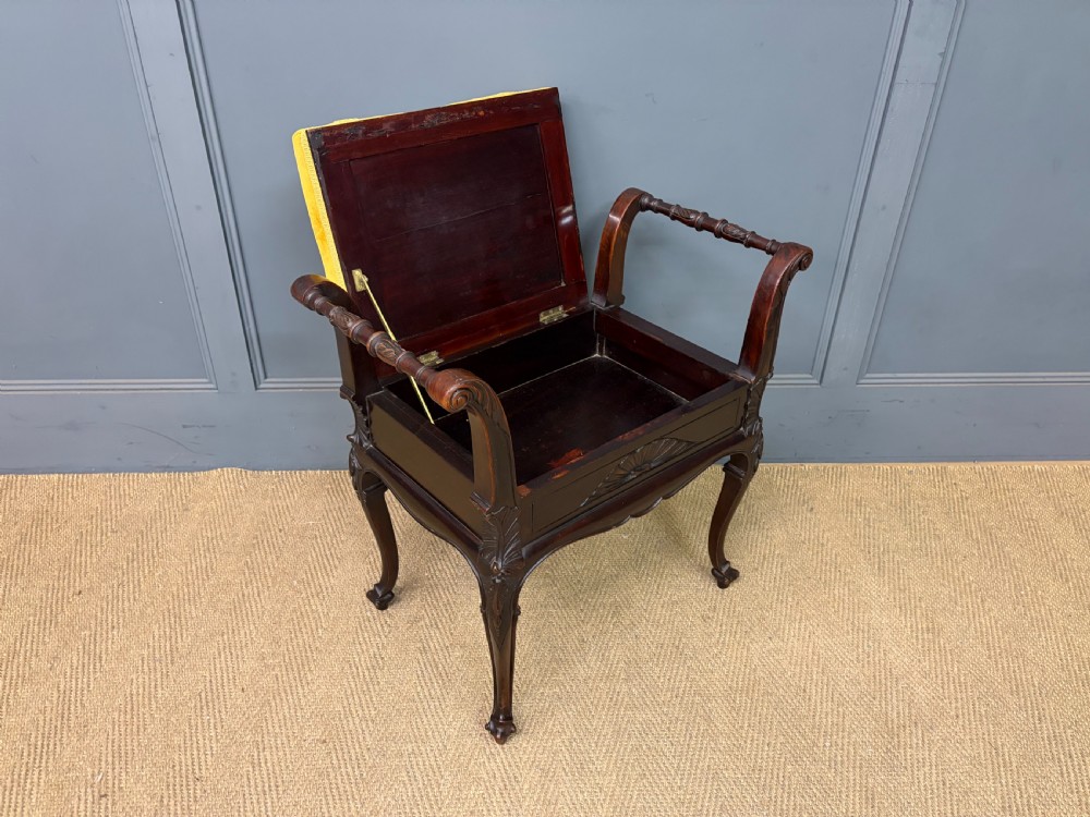 An antique dark mahogany piano stool featuring carved cabriole legs, scroll side handles, a carved shell apron motif, and a padded yellow velvet seat. View X