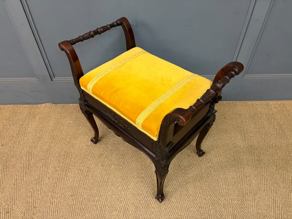 An antique dark mahogany piano stool featuring carved cabriole legs, scroll side handles, a carved shell apron motif, and a padded yellow velvet seat. View XII
