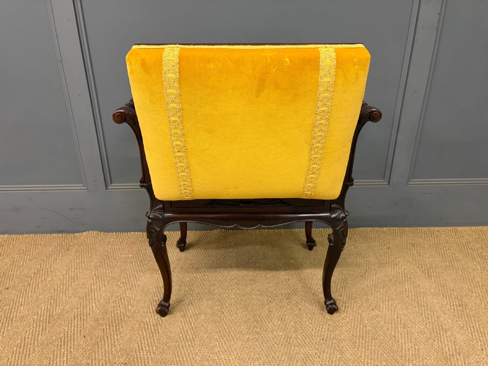 An antique dark mahogany piano stool featuring carved cabriole legs, scroll side handles, a carved shell apron motif, and a padded yellow velvet seat. View XV