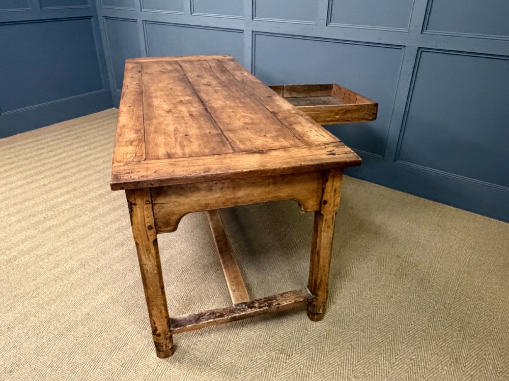 An early 19th-century fruitwood refectory table with a plank top, single side drawer, and stretcher base. View XXV