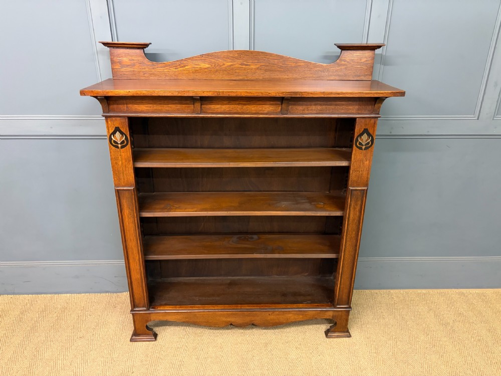 An Arts and Crafts medium brown oak open bookcase featuring four fixed shelves, a shaped top gallery, bracketed cornice, and stylised floral inlay at the top corners. View II