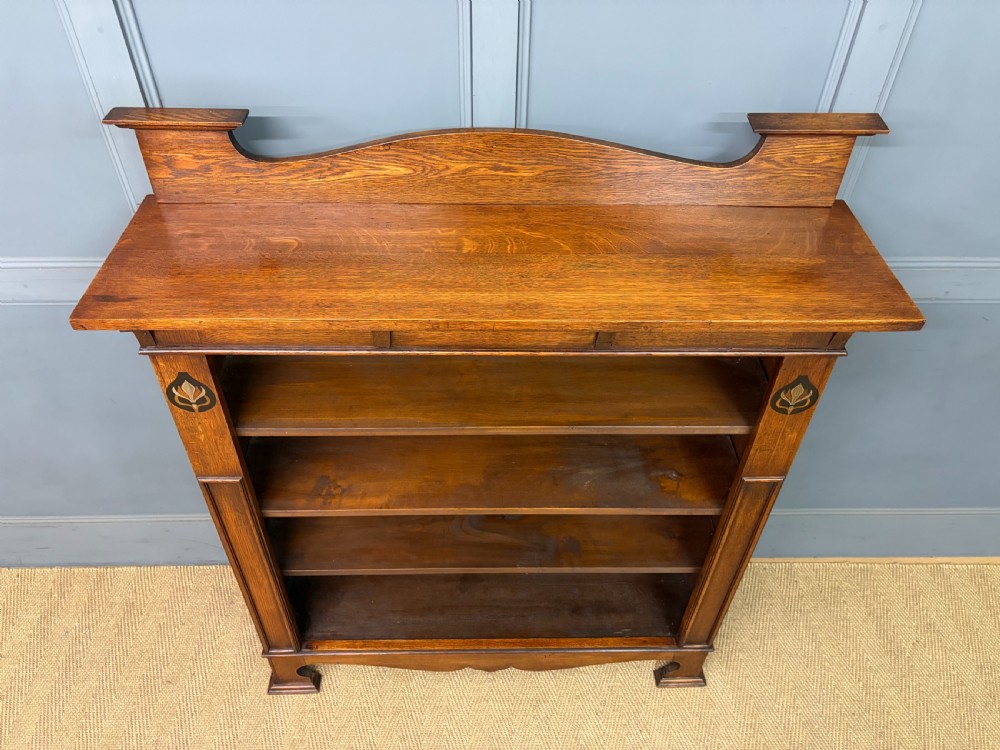 An Arts and Crafts medium brown oak open bookcase featuring four fixed shelves, a shaped top gallery, bracketed cornice, and stylised floral inlay at the top corners. View III