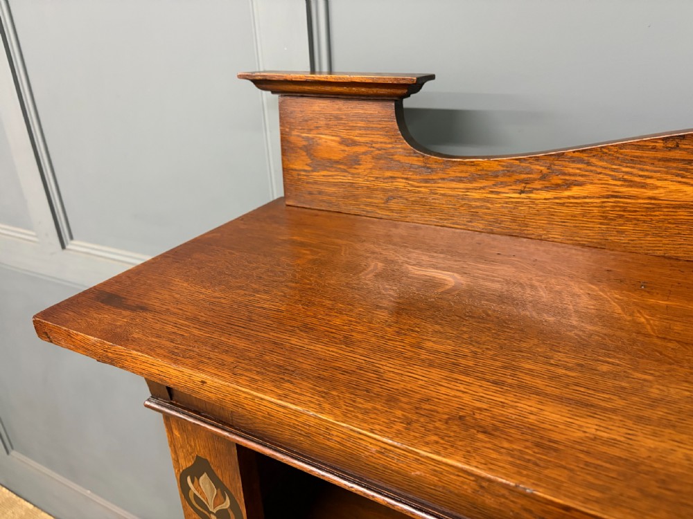 An Arts and Crafts medium brown oak open bookcase featuring four fixed shelves, a shaped top gallery, bracketed cornice, and stylised floral inlay at the top corners. View IV