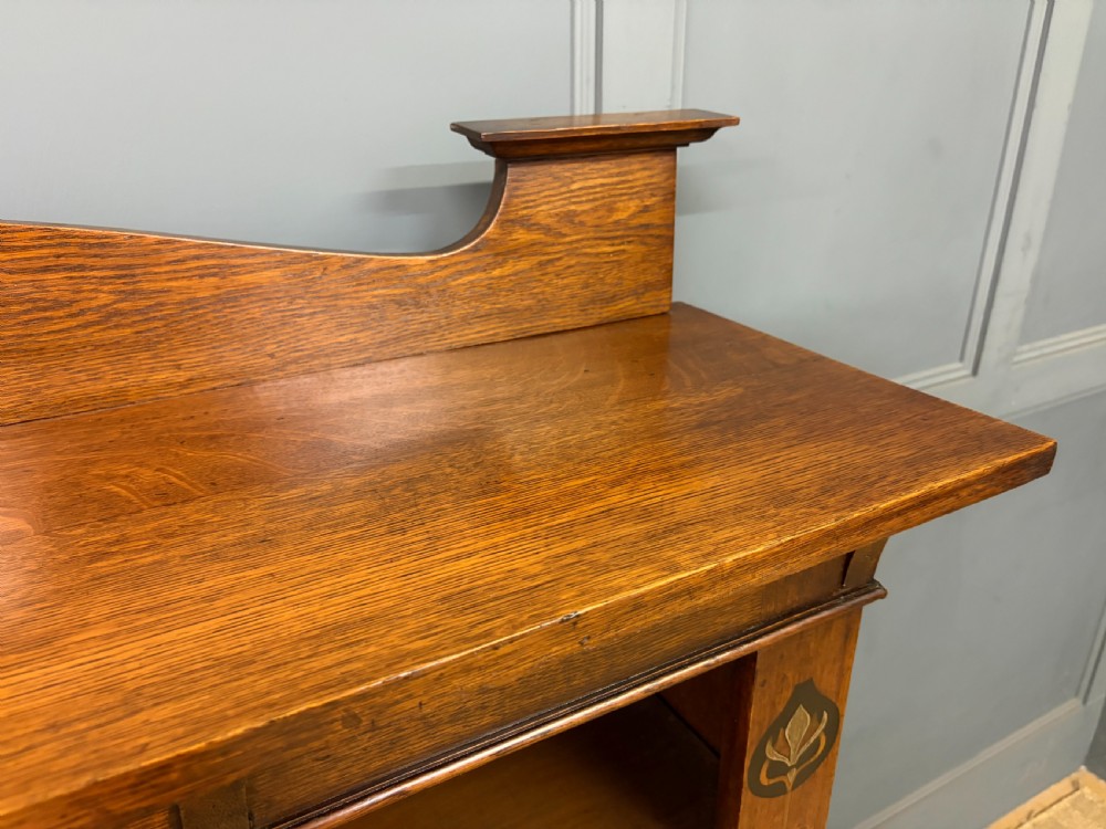 An Arts and Crafts medium brown oak open bookcase featuring four fixed shelves, a shaped top gallery, bracketed cornice, and stylised floral inlay at the top corners. View V