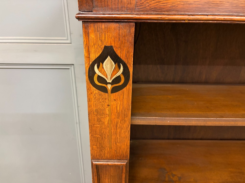 An Arts and Crafts medium brown oak open bookcase featuring four fixed shelves, a shaped top gallery, bracketed cornice, and stylised floral inlay at the top corners. View VI