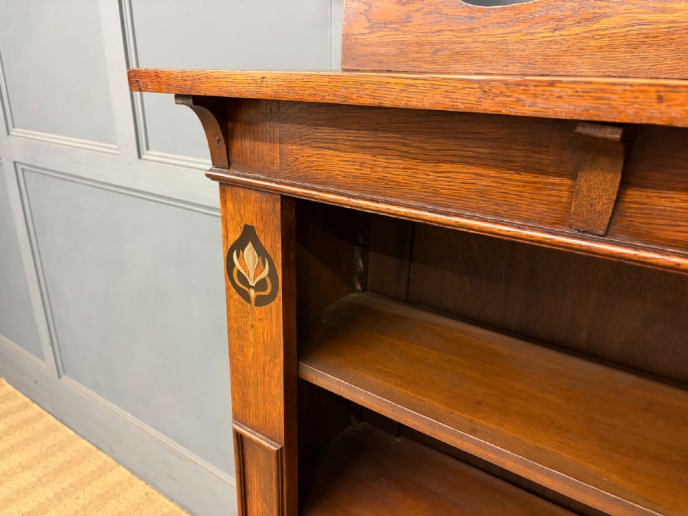 An Arts and Crafts medium brown oak open bookcase featuring four fixed shelves, a shaped top gallery, bracketed cornice, and stylised floral inlay at the top corners. View VII