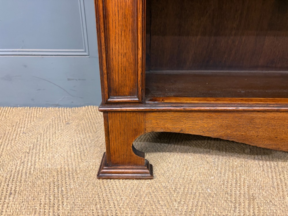 An Arts and Crafts medium brown oak open bookcase featuring four fixed shelves, a shaped top gallery, bracketed cornice, and stylised floral inlay at the top corners. View VIII
