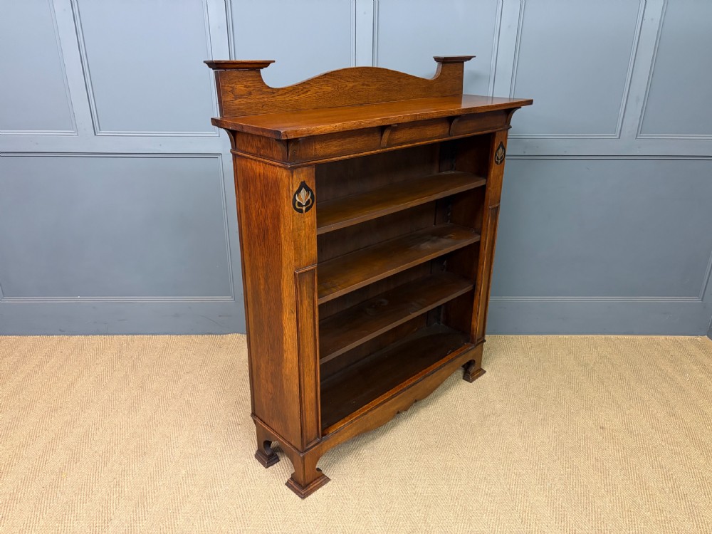 An Arts and Crafts medium brown oak open bookcase featuring four fixed shelves, a shaped top gallery, bracketed cornice, and stylised floral inlay at the top corners. View IX