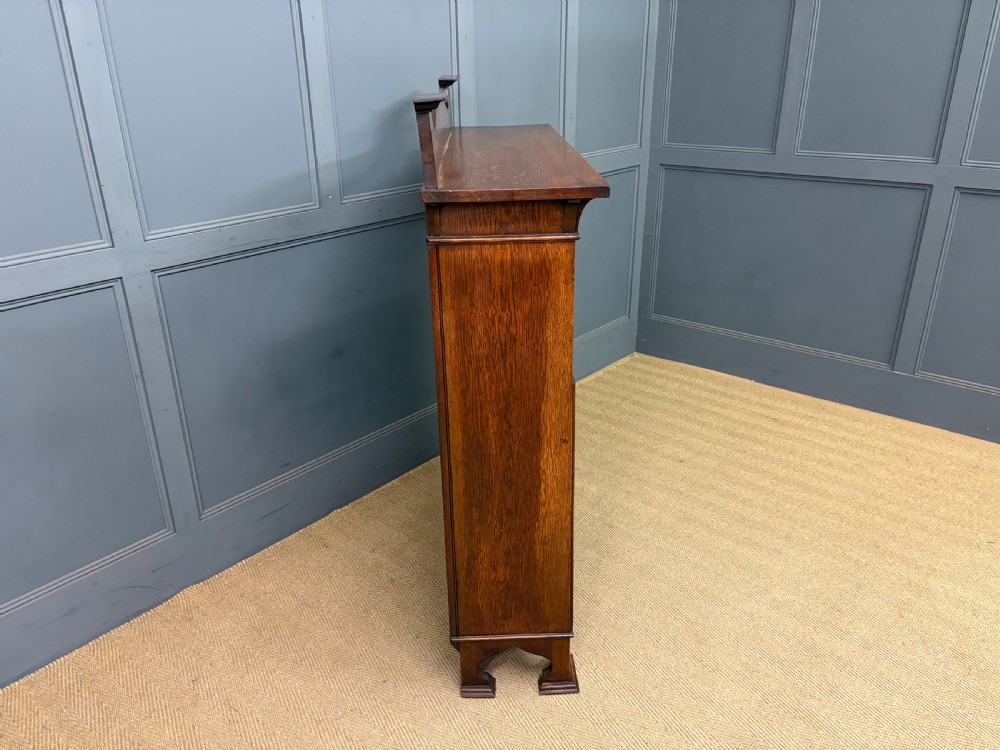 An Arts and Crafts medium brown oak open bookcase featuring four fixed shelves, a shaped top gallery, bracketed cornice, and stylised floral inlay at the top corners. View X