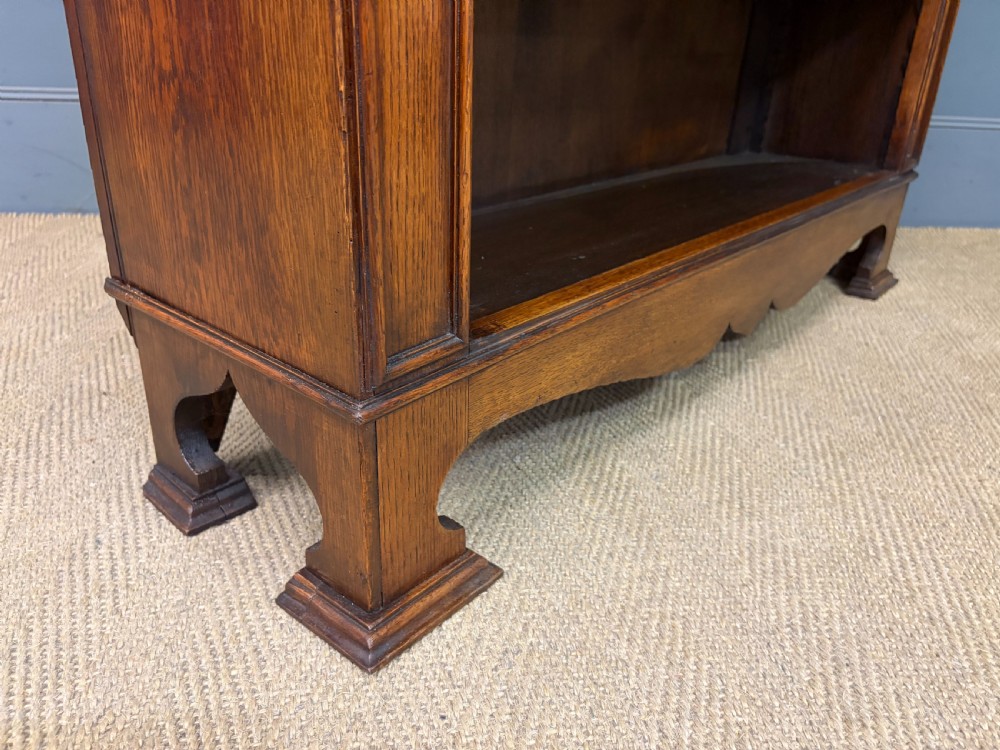 An Arts and Crafts medium brown oak open bookcase featuring four fixed shelves, a shaped top gallery, bracketed cornice, and stylised floral inlay at the top corners. View XI