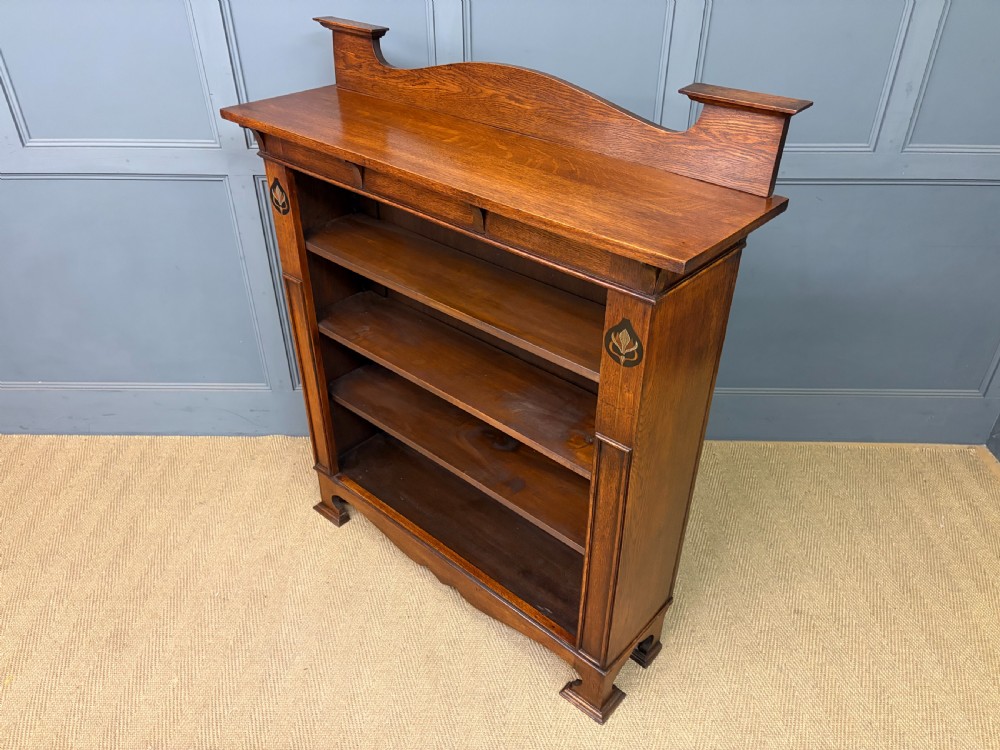 An Arts and Crafts medium brown oak open bookcase featuring four fixed shelves, a shaped top gallery, bracketed cornice, and stylised floral inlay at the top corners. View XII