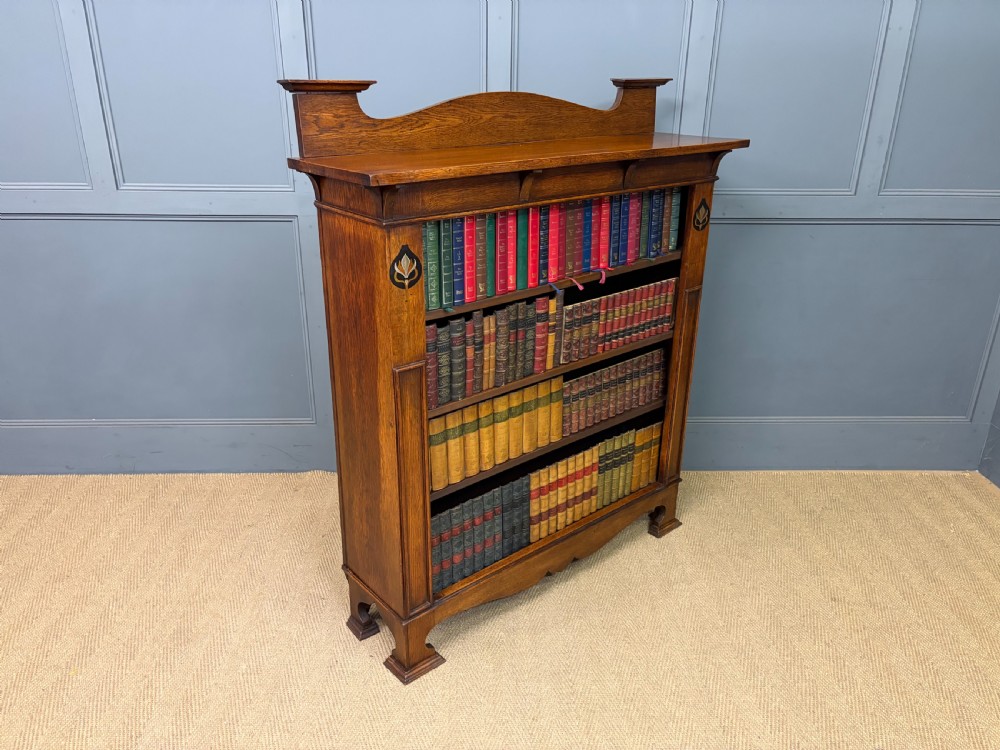 An Arts and Crafts medium brown oak open bookcase featuring four fixed shelves, a shaped top gallery, bracketed cornice, and stylised floral inlay at the top corners. View XIII