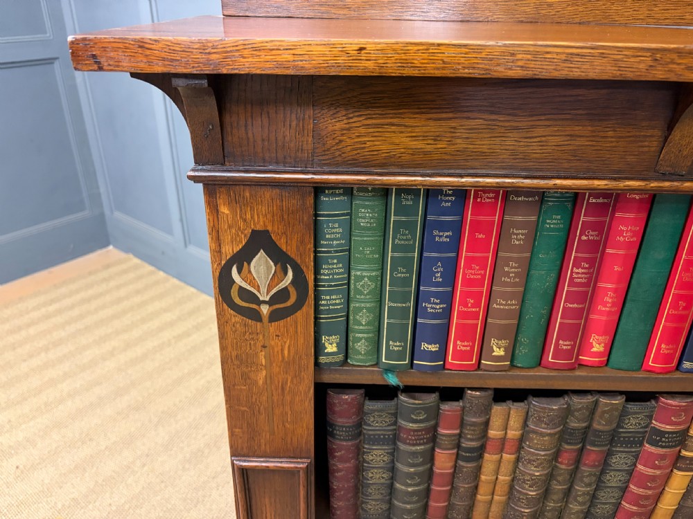 An Arts and Crafts medium brown oak open bookcase featuring four fixed shelves, a shaped top gallery, bracketed cornice, and stylised floral inlay at the top corners. View XIV