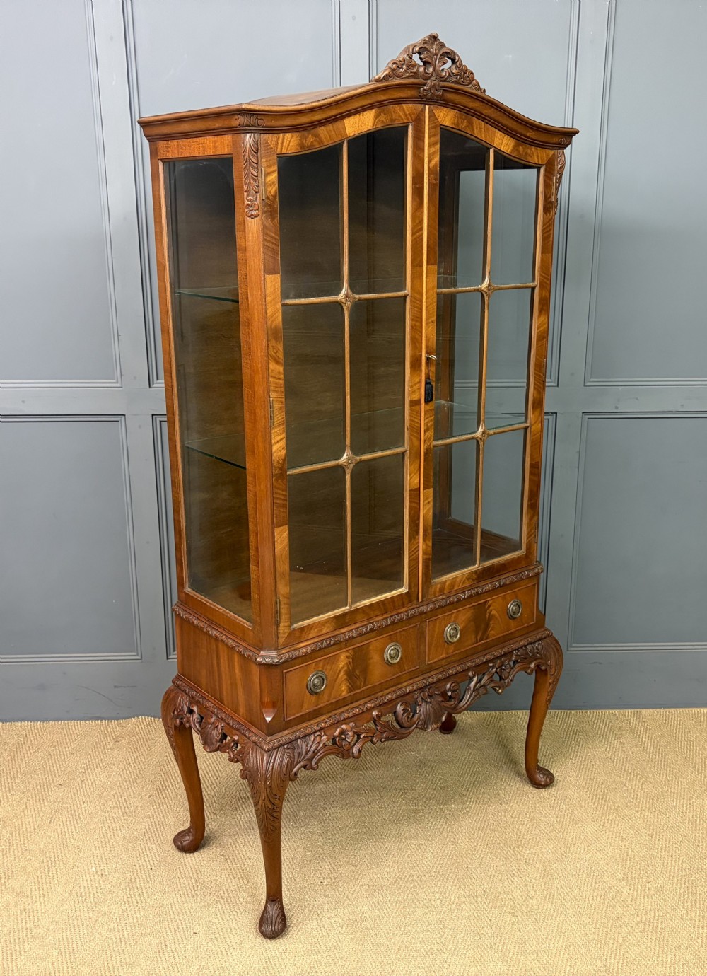 An antique Queen Anne style glazed display cabinet in warm brown wood featuring arched doors with glass panes, drawers with brass handles, and carved cabriole legs.