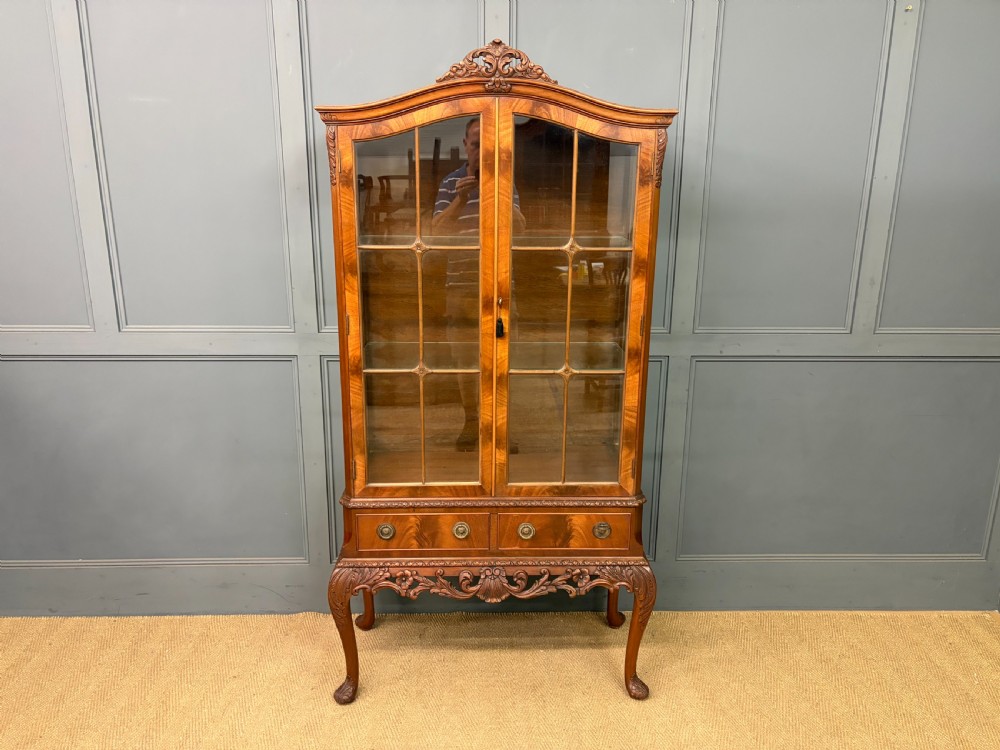 An antique Queen Anne style glazed display cabinet in warm brown wood featuring arched doors with glass panes, drawers with brass handles, and carved cabriole legs. View II