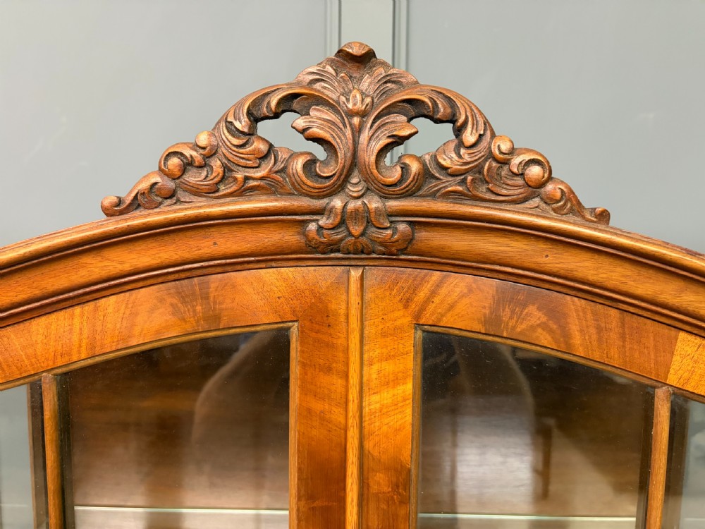 An antique Queen Anne style glazed display cabinet in warm brown wood featuring arched doors with glass panes, drawers with brass handles, and carved cabriole legs. View IV