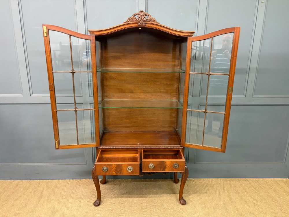 An antique Queen Anne style glazed display cabinet in warm brown wood featuring arched doors with glass panes, drawers with brass handles, and carved cabriole legs. View XI