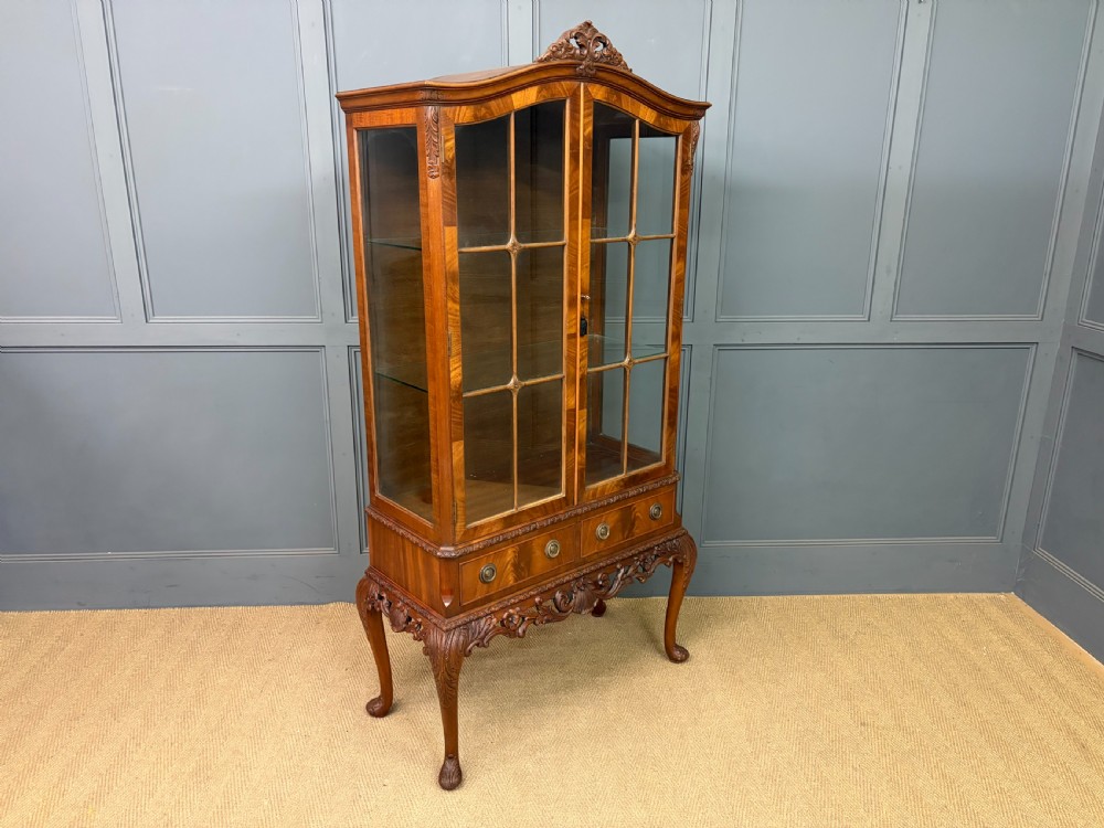 An antique Queen Anne style glazed display cabinet in warm brown wood featuring arched doors with glass panes, drawers with brass handles, and carved cabriole legs. View XIV