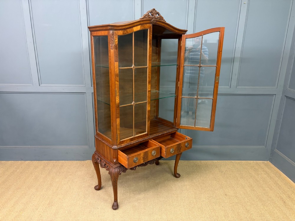 An antique Queen Anne style glazed display cabinet in warm brown wood featuring arched doors with glass panes, drawers with brass handles, and carved cabriole legs. View XIX