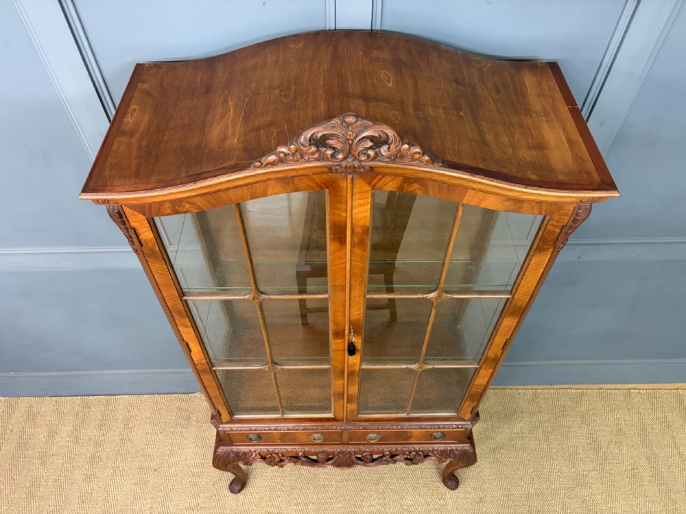An antique Queen Anne style glazed display cabinet in warm brown wood featuring arched doors with glass panes, drawers with brass handles, and carved cabriole legs. View XXII