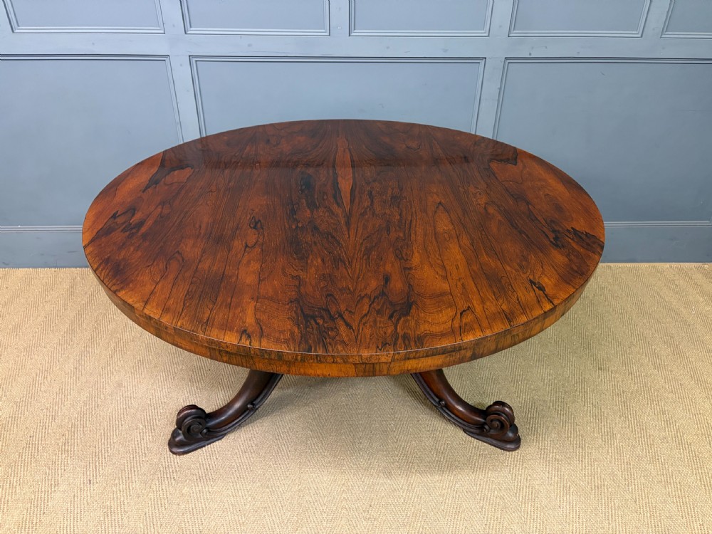 A Victorian oval rosewood loo table featuring a bookmatched veneer tabletop supported by a carved pedestal base with four scroll feet. View II
