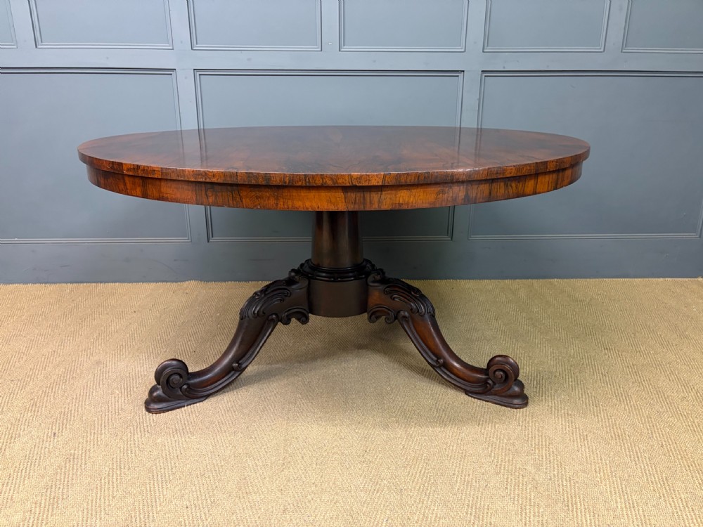A Victorian oval rosewood loo table featuring a bookmatched veneer tabletop supported by a carved pedestal base with four scroll feet. View III