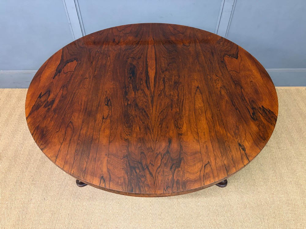 A Victorian oval rosewood loo table featuring a bookmatched veneer tabletop supported by a carved pedestal base with four scroll feet. View IV