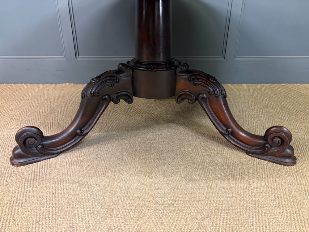 A Victorian oval rosewood loo table featuring a bookmatched veneer tabletop supported by a carved pedestal base with four scroll feet. View VI