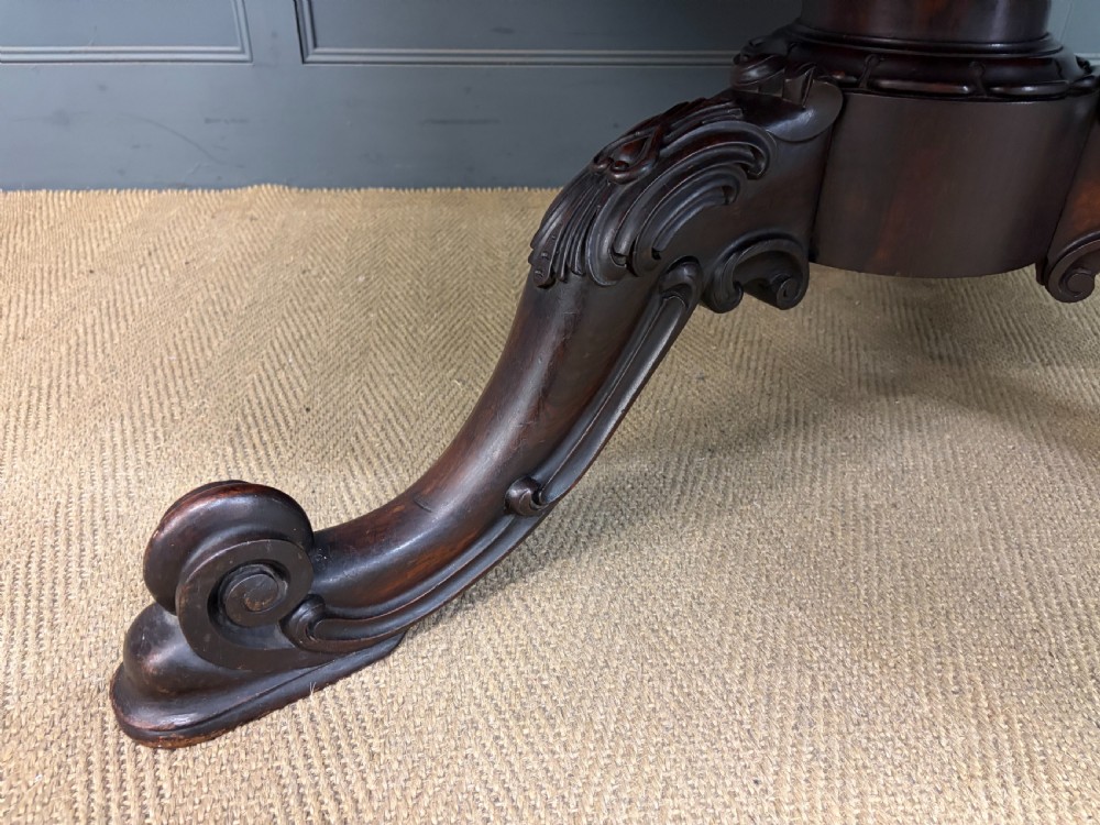 A Victorian oval rosewood loo table featuring a bookmatched veneer tabletop supported by a carved pedestal base with four scroll feet. View VII
