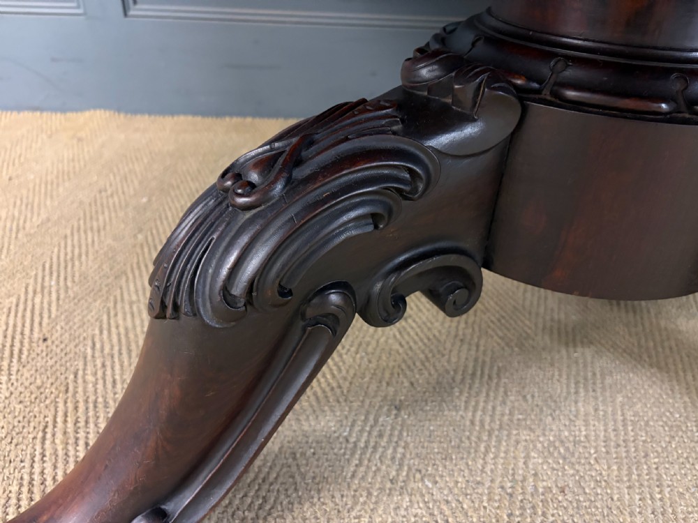 A Victorian oval rosewood loo table featuring a bookmatched veneer tabletop supported by a carved pedestal base with four scroll feet. View IX