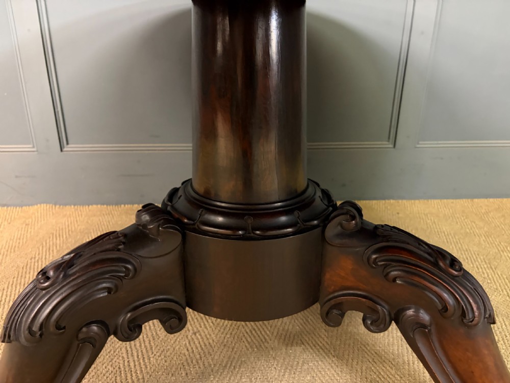 A Victorian oval rosewood loo table featuring a bookmatched veneer tabletop supported by a carved pedestal base with four scroll feet. View X