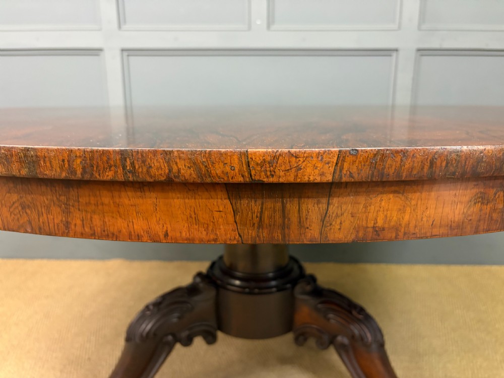 A Victorian oval rosewood loo table featuring a bookmatched veneer tabletop supported by a carved pedestal base with four scroll feet. View XI