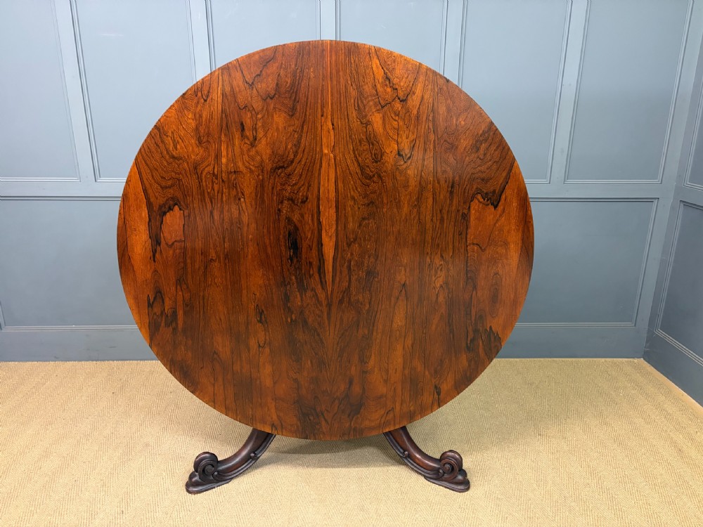 A Victorian oval rosewood loo table featuring a bookmatched veneer tabletop supported by a carved pedestal base with four scroll feet. View XII