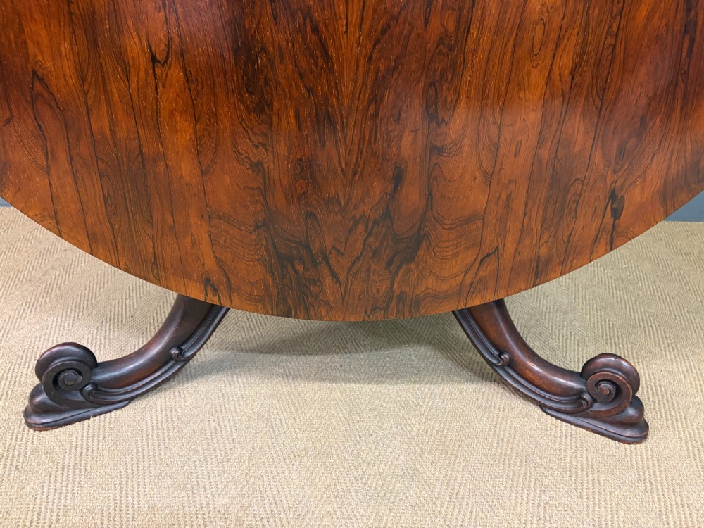 A Victorian oval rosewood loo table featuring a bookmatched veneer tabletop supported by a carved pedestal base with four scroll feet. View XIII