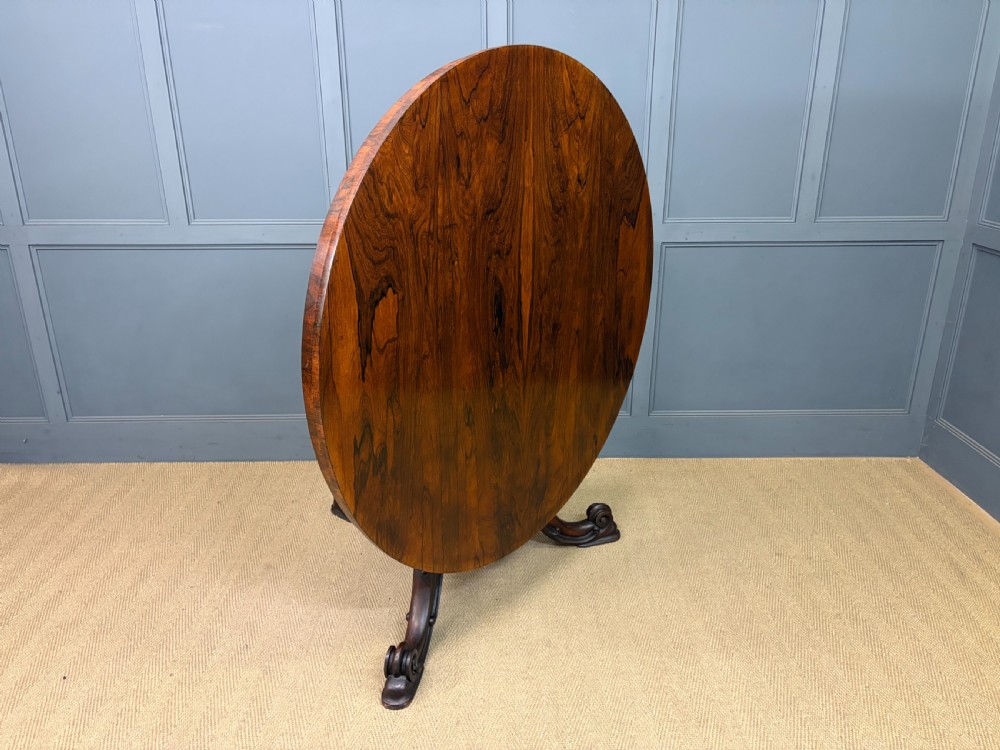 A Victorian oval rosewood loo table featuring a bookmatched veneer tabletop supported by a carved pedestal base with four scroll feet. View XV