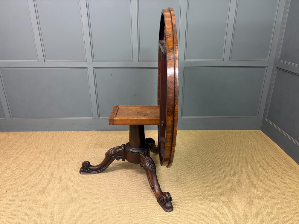 A Victorian oval rosewood loo table featuring a bookmatched veneer tabletop supported by a carved pedestal base with four scroll feet. View XVI