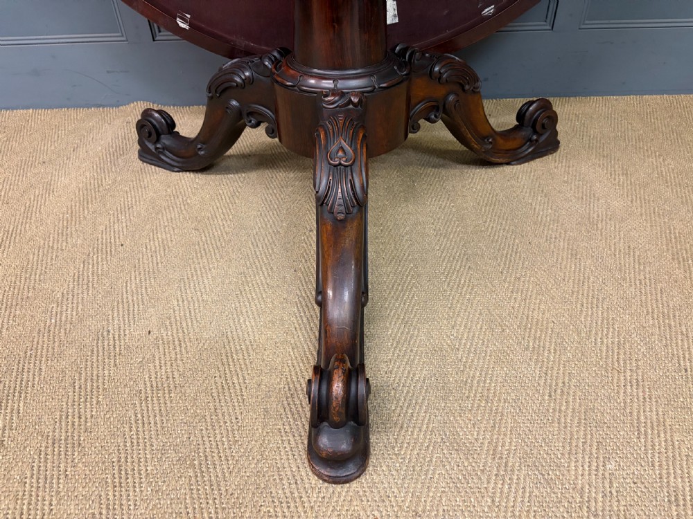 A Victorian oval rosewood loo table featuring a bookmatched veneer tabletop supported by a carved pedestal base with four scroll feet. View XVIII
