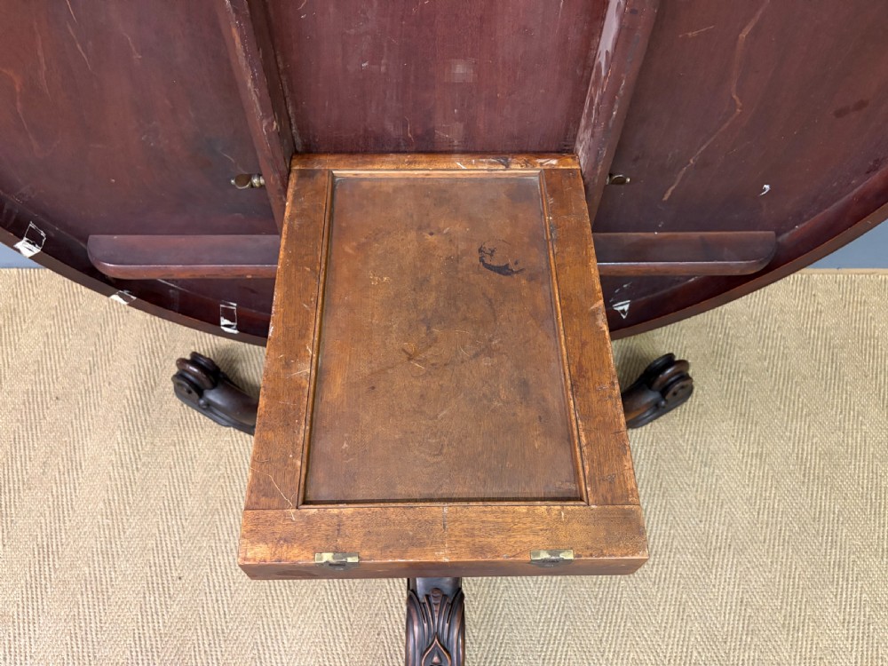 A Victorian oval rosewood loo table featuring a bookmatched veneer tabletop supported by a carved pedestal base with four scroll feet. View XX