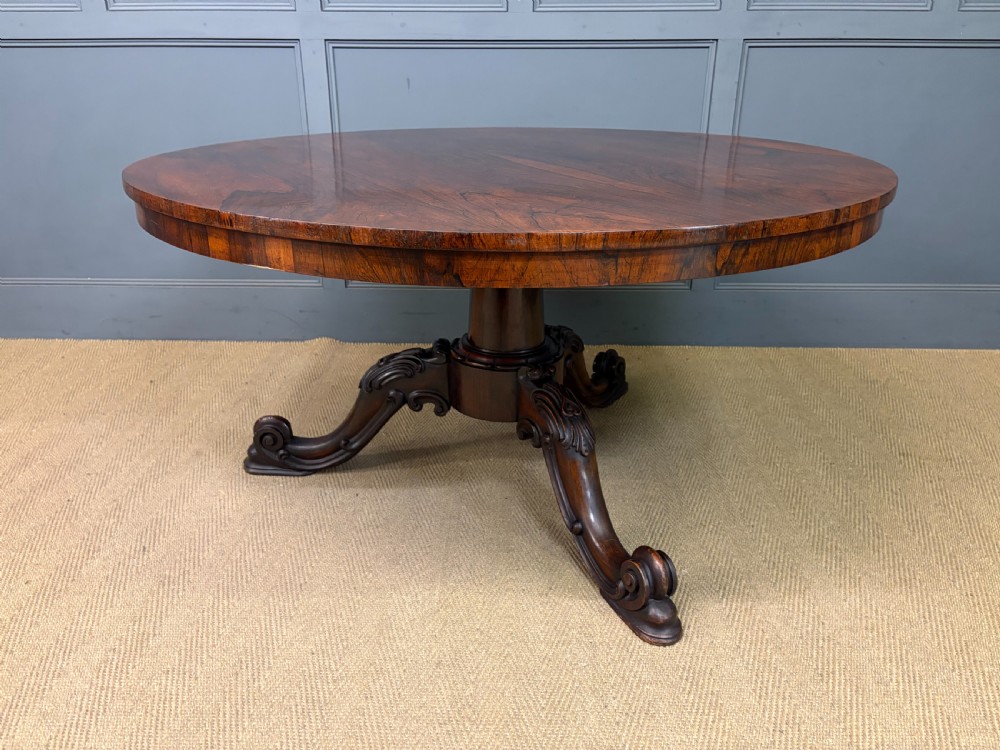 A Victorian oval rosewood loo table featuring a bookmatched veneer tabletop supported by a carved pedestal base with four scroll feet. View XXI